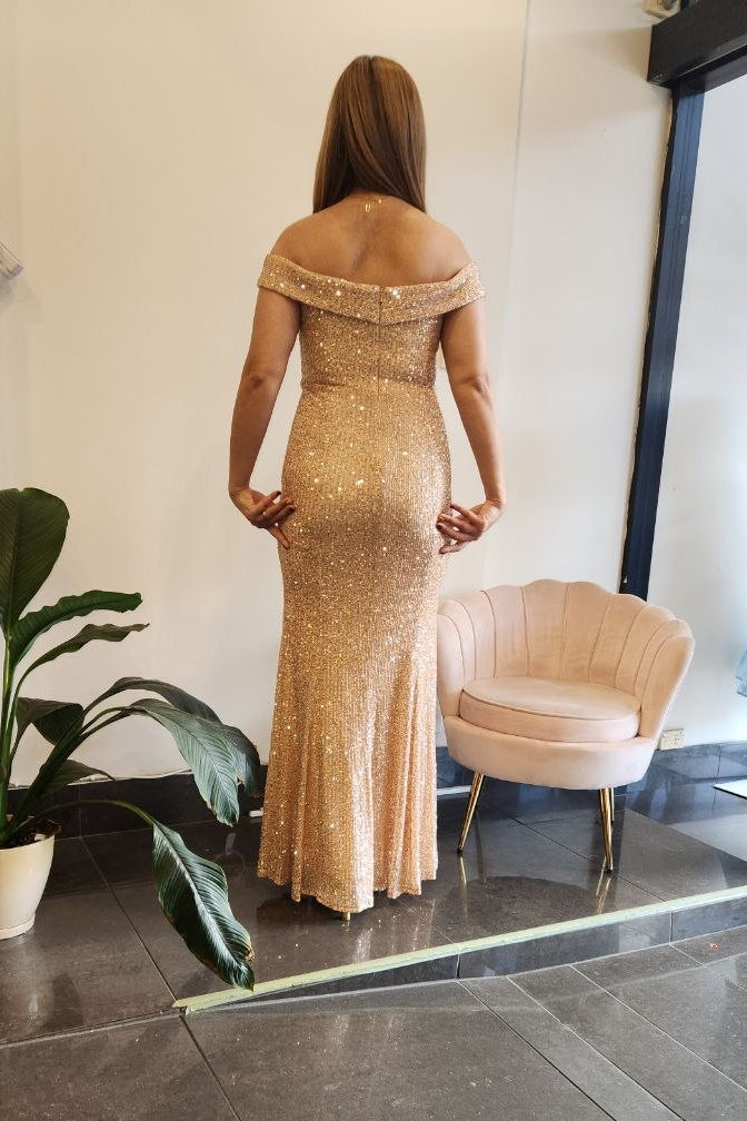 A woman stands with her back to the camera wearing a floor-length off-the-shoulder champagne gold sequined gown in a boutique showroom with a pink chair and a potted plant.