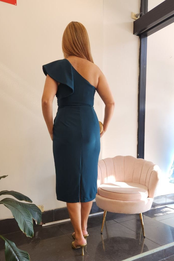 A woman stands with her back to the camera wearing a dark teal one-shoulder midi dress in a boutique setting with a pink chair and a mannequin displaying a blue dress in the background.