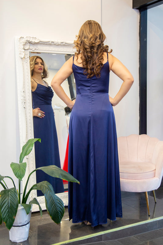 Woman in a blue dress standing in front of a mirror in a room with a plant and chair.