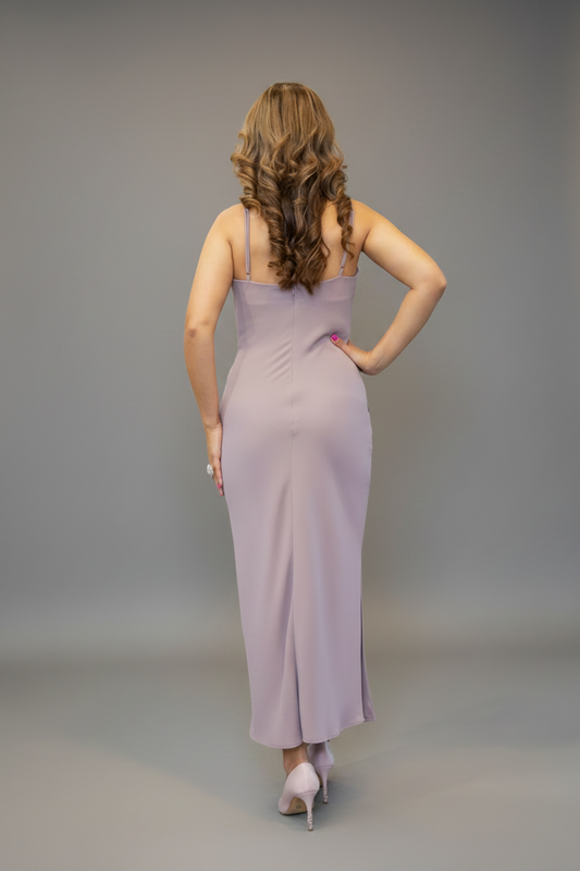 A woman with long wavy brown hair is shown from the back wearing a lavender sleeveless maxi dress with thin straps and a side slit, paired with white heels, against a light gray studio background.