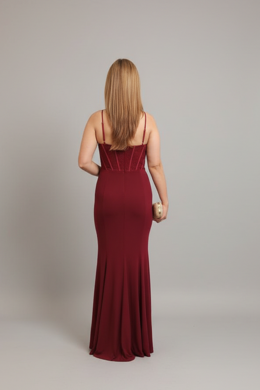 A woman viewed from behind wearing a floor-length burgundy maxi gown with thin straps and structured detailing at the back, standing against a plain light gray studio background.