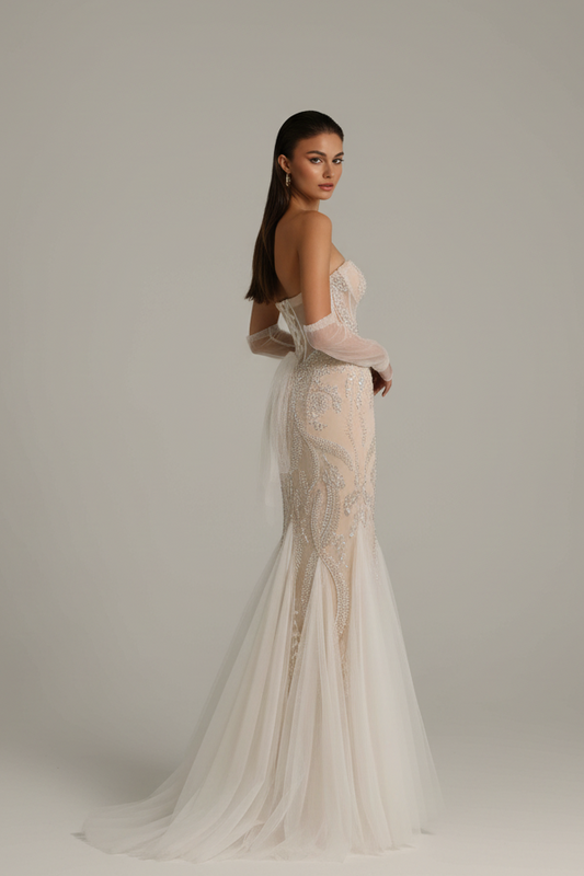 A woman modeling a strapless off-white mermaid-style wedding gown with sheer long sleeves and a flowing tulle train, photographed against a plain light gray studio background.