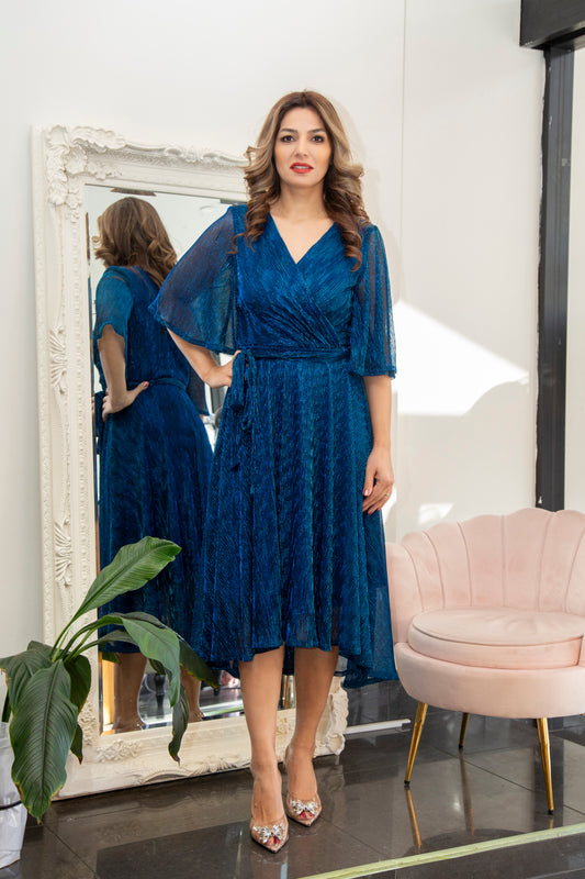 Woman in a blue dress standing in front of a mirror in a room with a pink chair and plant.