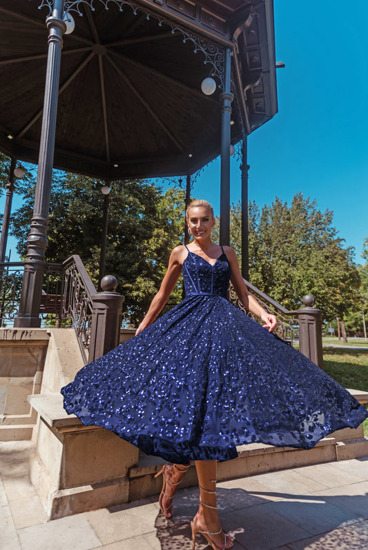 Woman in a blue sequin dress standing in front of a gazebo with trees and clear sky in the background.