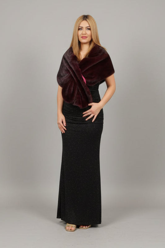 A woman stands indoors wearing a dark plum wrap-style top and a long black skirt. The setting appears to be a boutique or showroom with a pale pink chair, a large potted plant, tiled floor, and glass door.