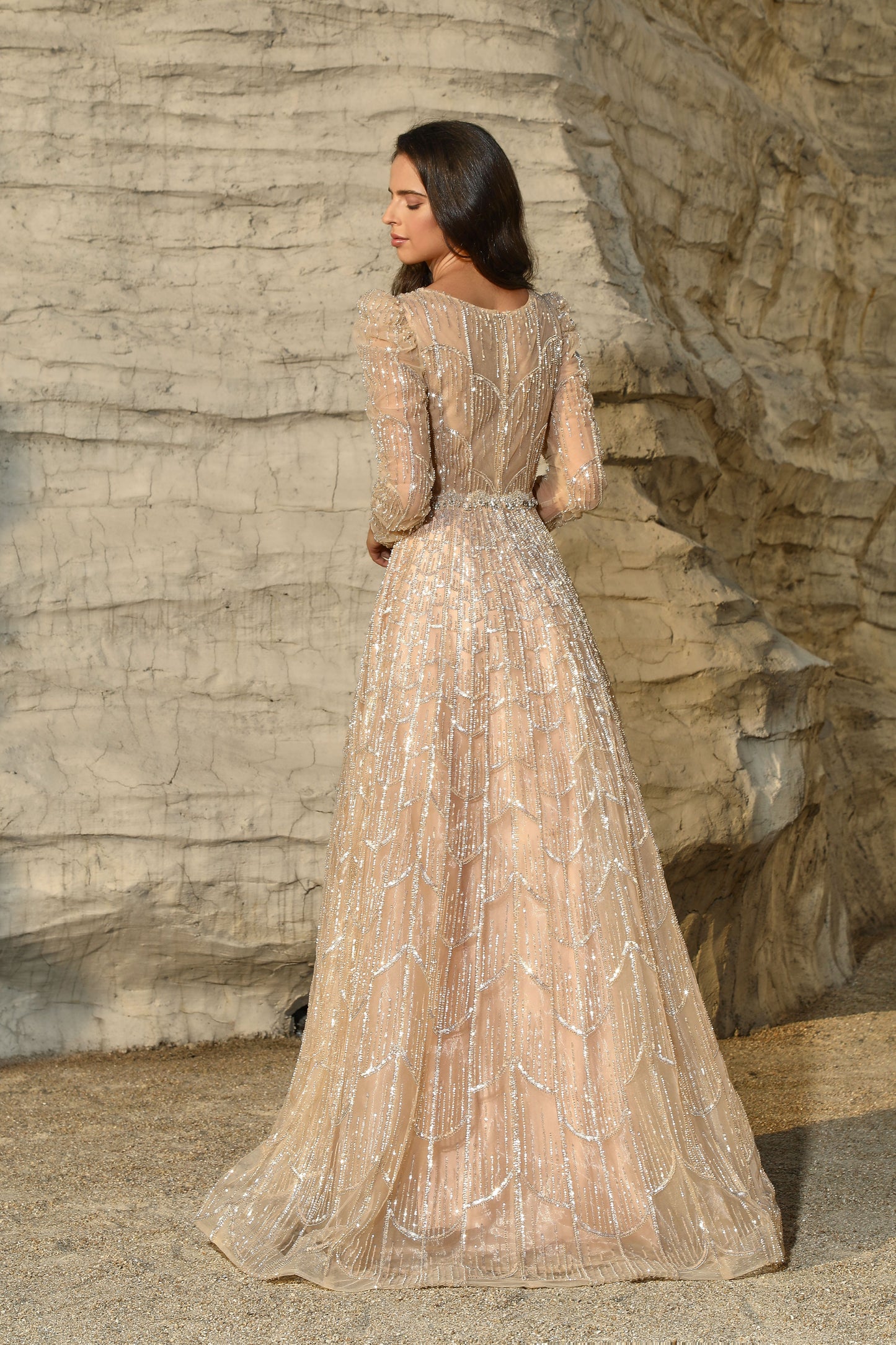 Woman in a beige evening gown standing against a stone wall.