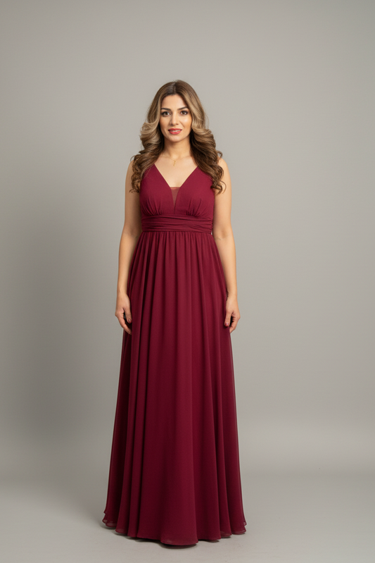 A woman modeling a sleeveless, floor-length burgundy formal gown with a V-neckline and gathered waist, standing against a neutral gray studio backdrop.