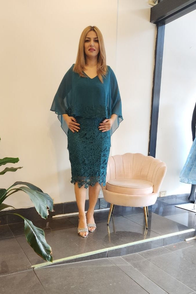 A woman modeling a teal blue-green lace midi dress with sheer flutter sleeves, standing in a boutique next to a pink shell chair.
