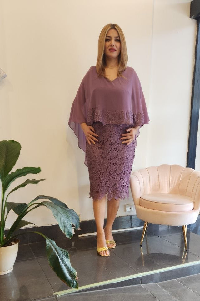 A woman models a matching mauve purple two-piece set consisting of a loose short-sleeve top and a textured lace midi skirt, standing in a boutique showroom with a pink chair and a potted plant.
