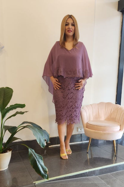 A woman models a matching mauve purple two-piece set consisting of a loose short-sleeve top and a textured lace midi skirt, standing in a boutique showroom with a pink chair and a potted plant.