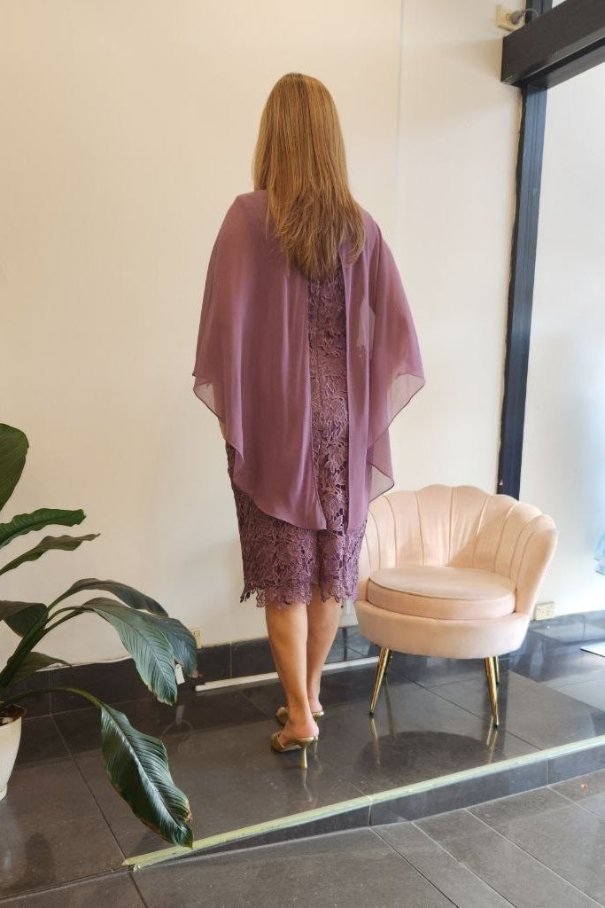 A woman stands with her back to the camera wearing a sheer mauve purple lace dress with a matching open-front shawl. She is posing in a boutique interior with a tiled floor, a potted plant, and a pink shell-shaped chair.