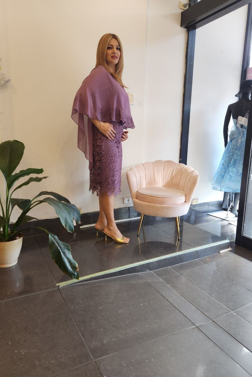 A woman models a lavender purple lace dress with a matching draped shawl and gold heels in a boutique setting, standing next to a pink velvet accent chair.