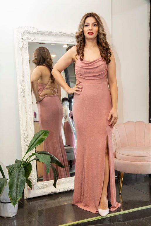 Woman in a pink dress standing in front of a mirror in a room with a plant and chair.