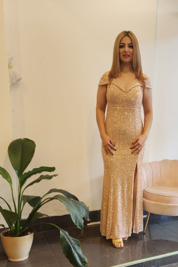 A woman stands indoors wearing a floor-length champagne beige sequined gown with cap sleeves and a front slit. The setting appears to be a boutique or showroom with a potted plant, a pale pink chair, dark tiled floor, and a mannequin in a blue dress visible through a glass door.