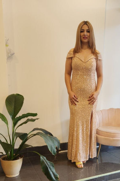 A woman stands indoors wearing a floor-length champagne beige sequined gown with cap sleeves and a front slit. The setting appears to be a boutique or showroom with a potted plant, a pale pink chair, dark tiled floor, and a mannequin in a blue dress visible through a glass door.