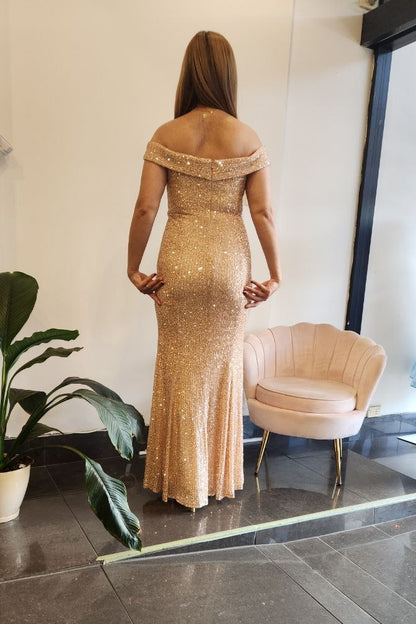 A woman stands with her back to the camera wearing a floor-length off-the-shoulder champagne gold sequined gown in a boutique showroom with a pink chair and a potted plant.