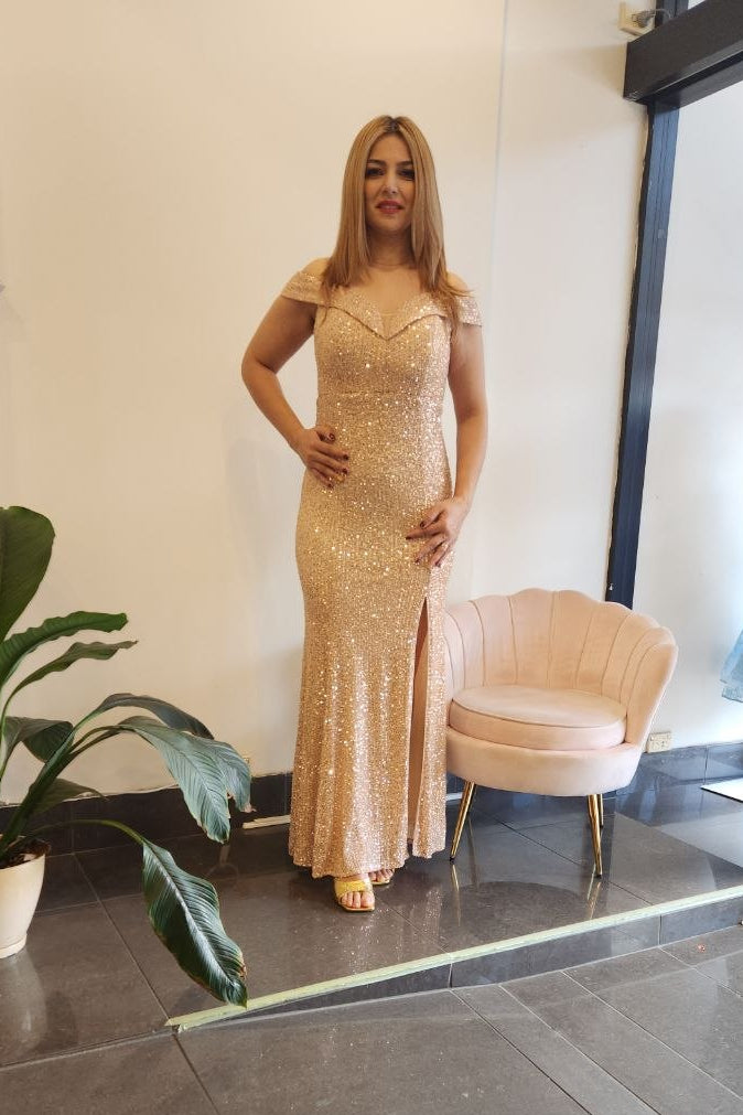 A woman modeling a champagne gold sequined floor-length evening gown with a sweetheart neckline and off-the-shoulder straps, standing in a boutique interior next to a pink shell chair and a potted plant.