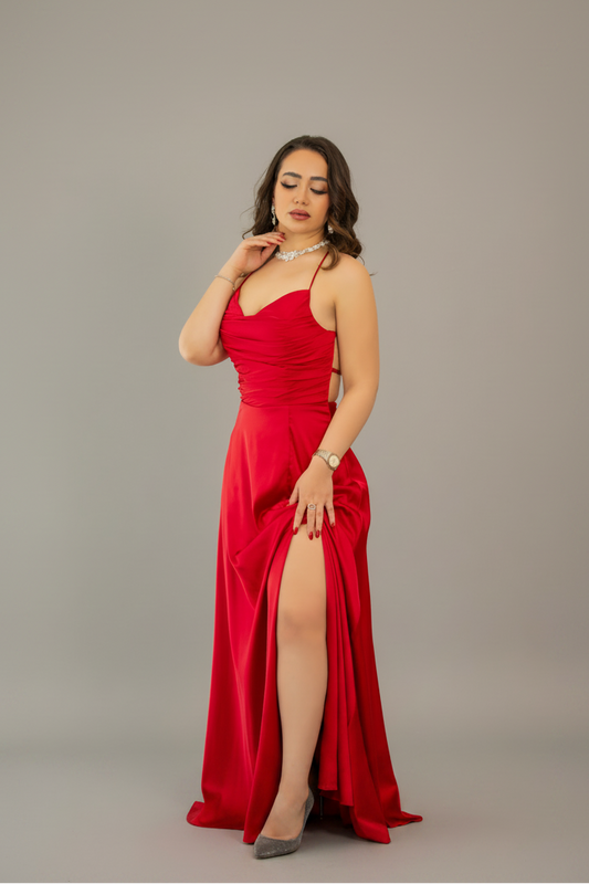 A woman modeling a floor-length red evening gown with thin straps, a sweetheart neckline, ruched detailing, and a high thigh slit, photographed against a plain gray studio background.