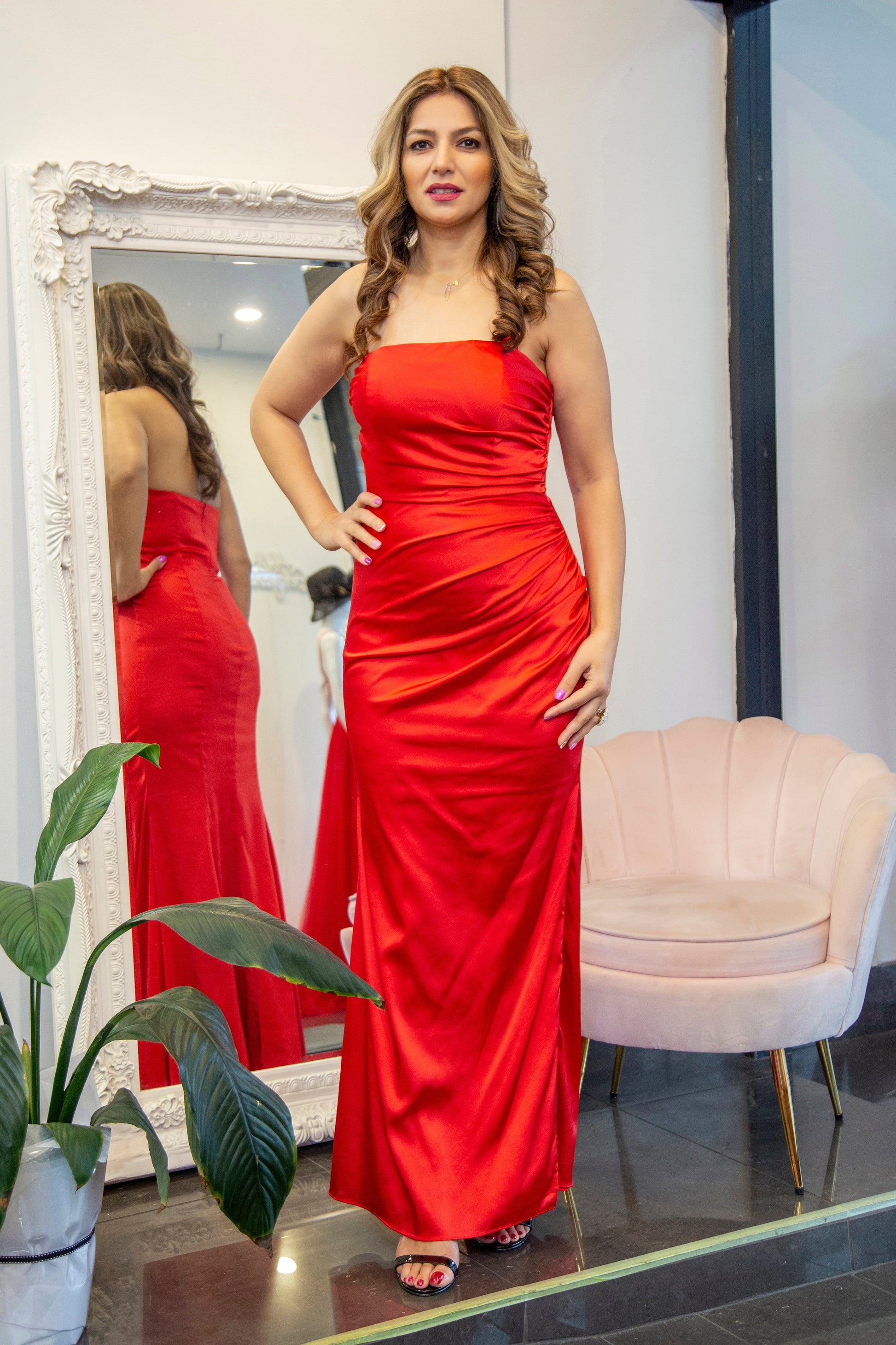 Woman in a red dress standing in front of a mirror in a room with a chair and plant.