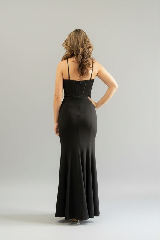 A woman stands with her back to the camera wearing a floor-length black spaghetti-strap gown in a minimalist studio setting with a light gray background.