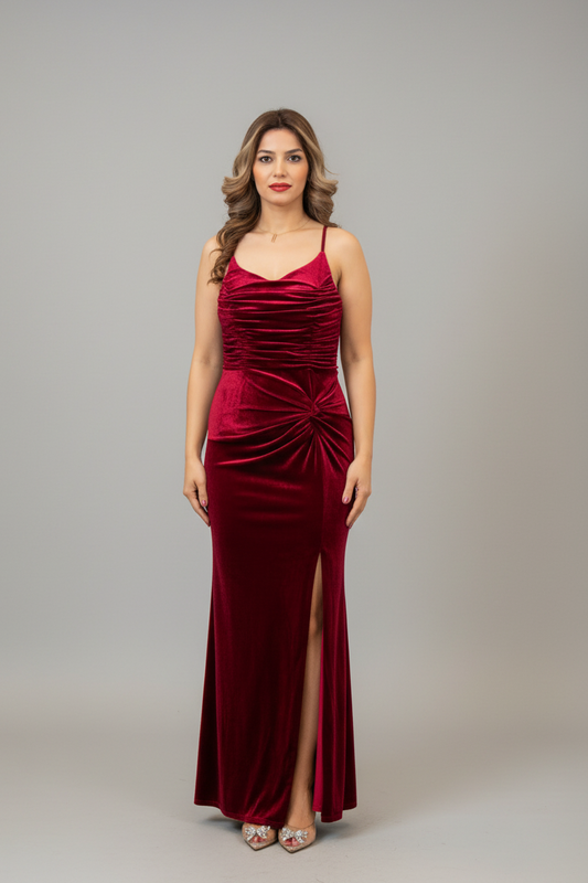 A woman modeling a floor-length burgundy velvet gown with spaghetti straps, a sweetheart neckline, ruched detailing, and a high thigh slit, photographed against a neutral gray studio background.