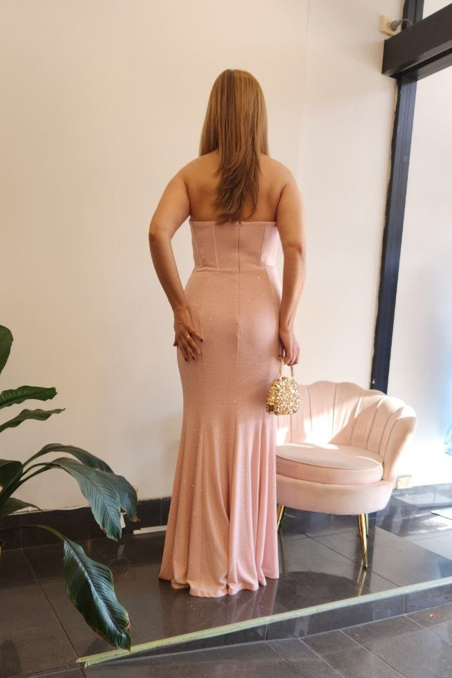 A woman stands with her back to the camera wearing a strapless blush pink floor-length gown in a boutique. A small pink velvet chair, potted plant, and glass door are visible in the store interior.