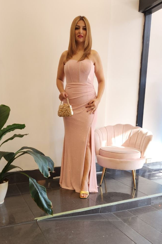 A woman stands indoors wearing a strapless, floor-length blush pink gown with a fitted bodice and slight sheen. She holds a small metallic handbag and wears gold heels.