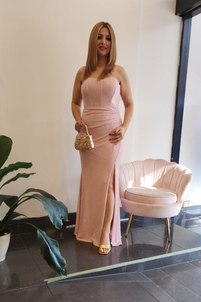 A woman stands indoors wearing a sleeveless, floor-length blush pink gown with a fitted bodice and draped skirt. She is holding a small metallic handbag beside a pale pink upholstered chair, with a large potted plant and a window view in the background.