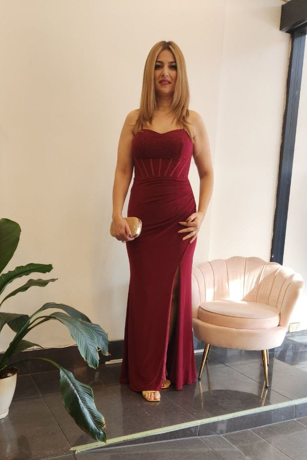 A woman modeling a sleeveless, deep burgundy red floor-length gown with a sweetheart neckline and a thigh-high front slit, standing in a boutique showroom with a pink shell chair and a potted plant nearby.