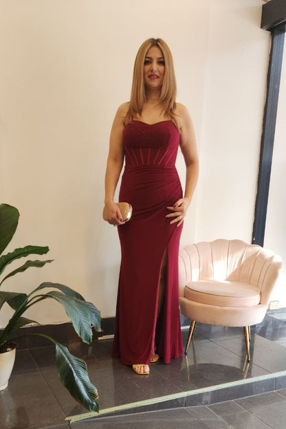 A woman modeling a sleeveless, deep burgundy red floor-length gown with a sweetheart neckline and a thigh-high front slit, standing in a boutique showroom with a pink shell chair and a potted plant nearby.