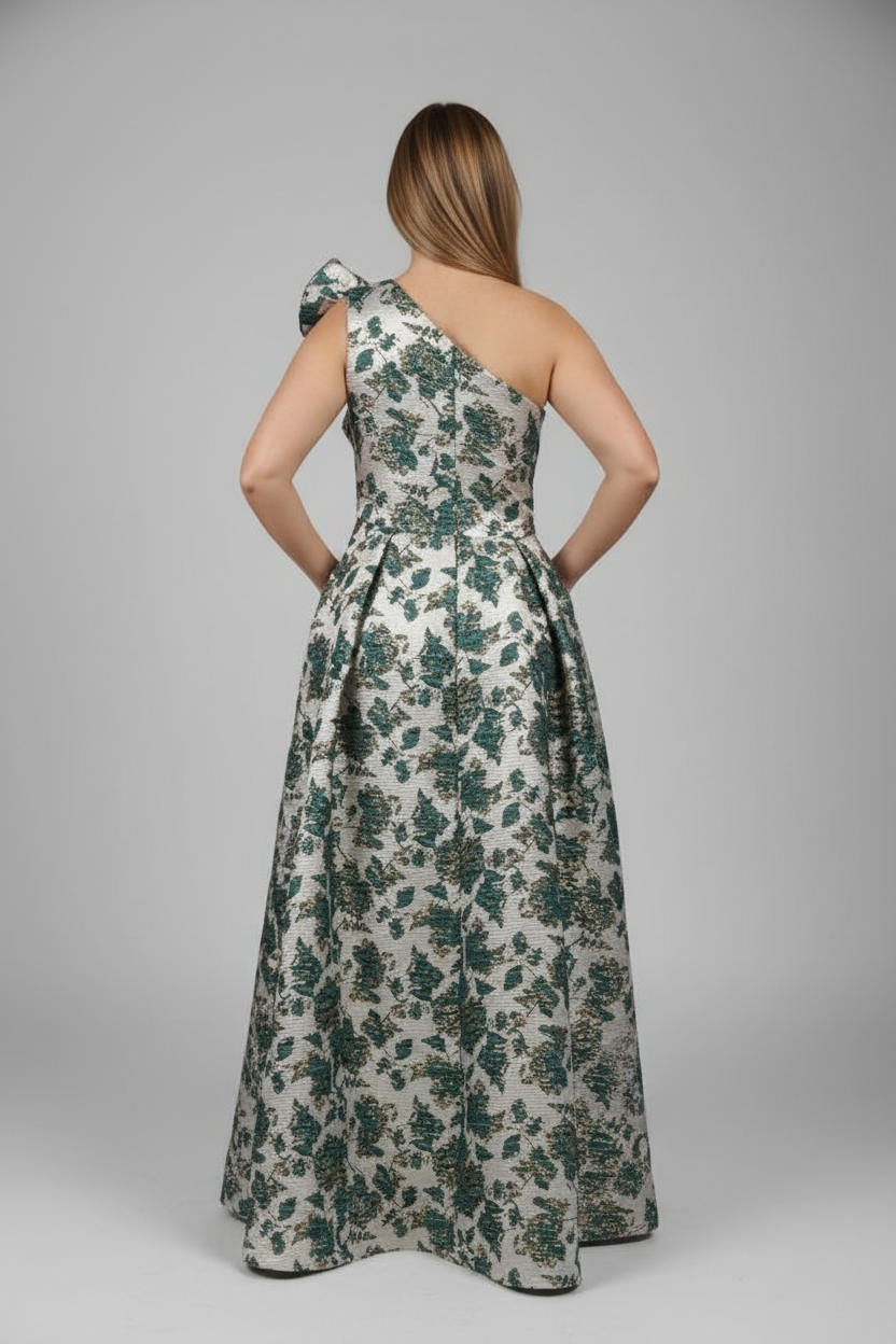 A woman stands with her back to the camera wearing a sleeveless, one-shoulder maxi dress with a floral print. The dress has a cream base with green and dark blue floral motifs.