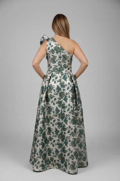 A woman stands with her back to the camera wearing a sleeveless, one-shoulder maxi dress with a floral print. The dress has a cream base with green and dark blue floral motifs.