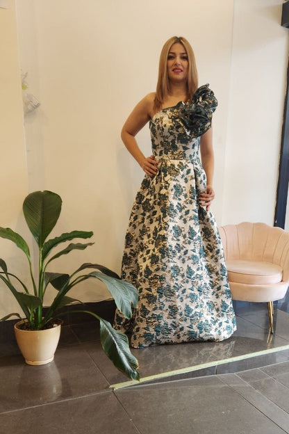 A woman models a strapless floor-length dress with a floral print in green, blue, and white, standing in a boutique interior with a pink chair and a potted plant.