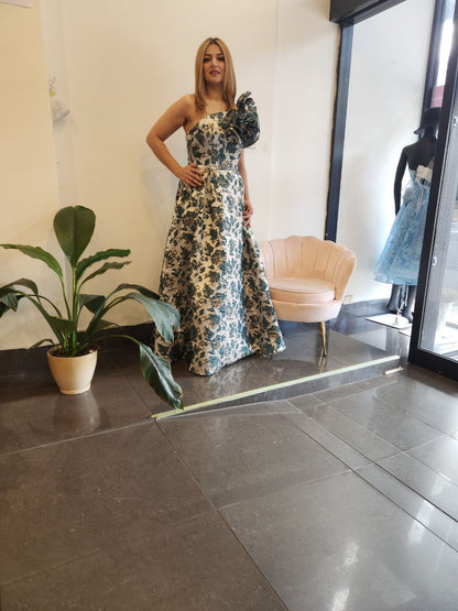 A woman models a strapless maxi dress with a ruffled sleeve and a blue, green, and beige floral pattern, standing in a boutique interior with a pink chair and a potted plant.