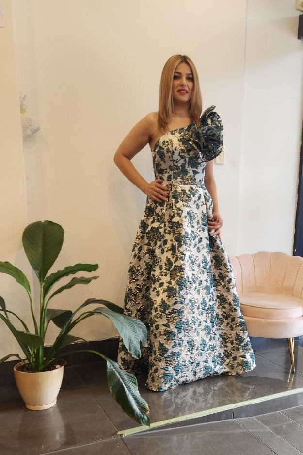 A woman models a strapless floor-length gown with a floral pattern in blue, green, and beige, featuring a ruffled one-shoulder detail. She is standing in a boutique interior with a potted plant, a pink chair, and a window display in the background.
