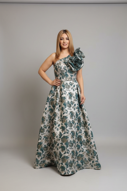 A woman modeling a floor-length one-shoulder gown with a ruffled shoulder detail and an allover floral pattern in dark teal-green and cream, set against a plain light gray studio background.