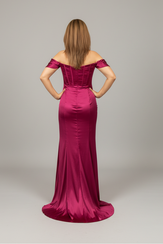 A woman viewed from the back wearing a floor-length off-the-shoulder gown in a rich magenta satin-like fabric, standing against a plain light gray studio background.