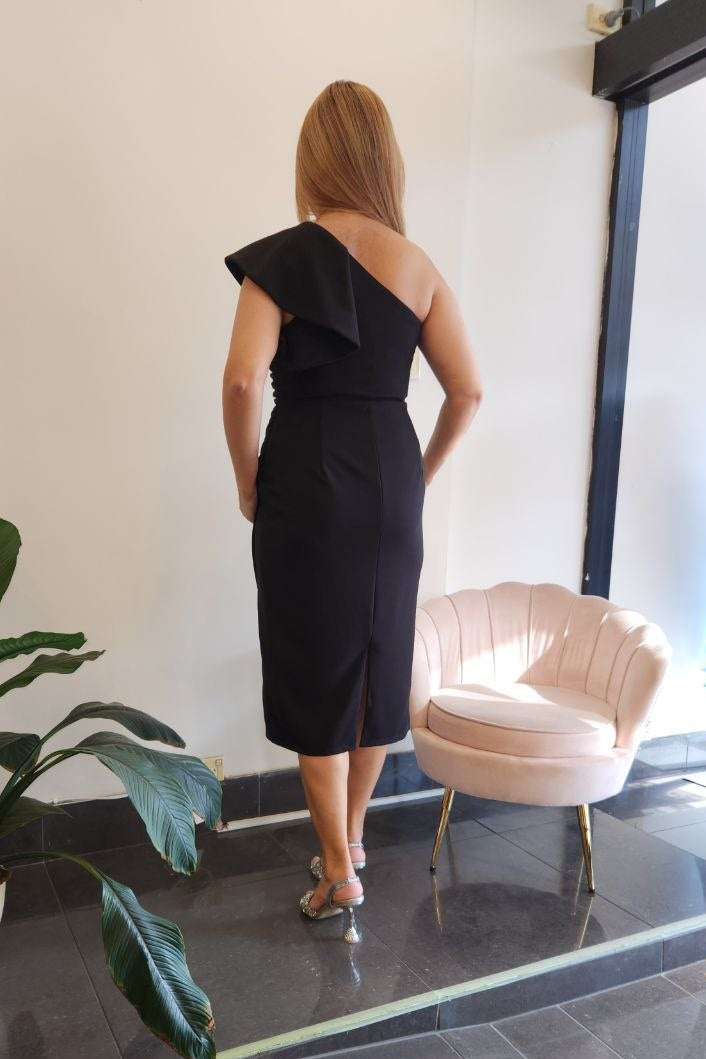 A woman stands with her back to the camera wearing a black one-shoulder midi dress in a boutique setting with a pink chair and a potted plant.