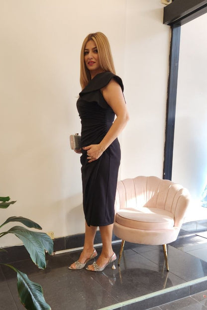 A woman wearing a black ruffled sleeveless dress and clear high-heeled sandals stands in a boutique interior next to a pink shell chair and a potted plant.