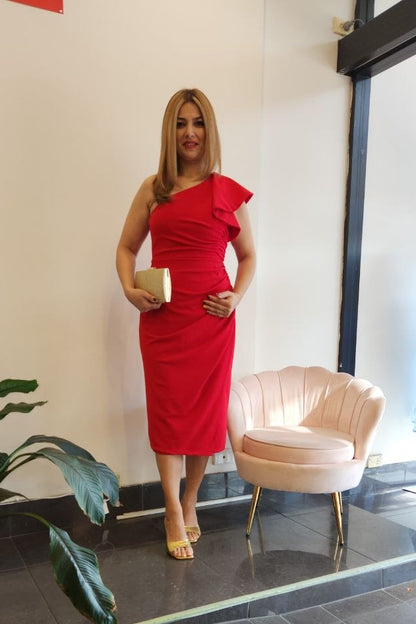 A woman modeling a bright red one-shoulder midi dress with a ruffled shoulder detail, standing in a boutique interior next to a pink chair and a potted plant.