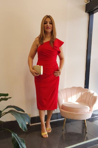 A woman stands indoors wearing a bright red one-shoulder midi dress with a ruffled shoulder detail and holding a small beige clutch. The setting includes a pale pink shell-shaped chair, a potted plant, and a window display with a mannequin wearing a blue dress.