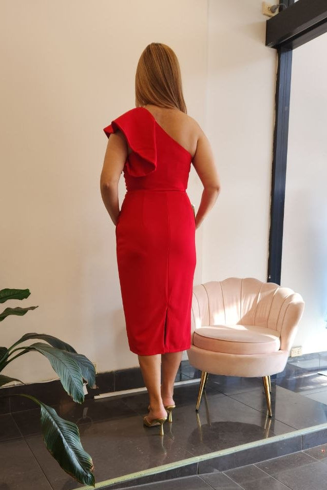 A woman stands with her back to the camera wearing a bright red one-shoulder midi dress in a boutique interior with a pink chair, tiled floor, and a mannequin displaying a blue dress in the window.