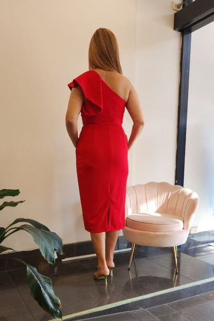 A woman stands with her back to the camera wearing a bright red one-shoulder midi dress in a boutique interior with a pink chair, tiled floor, and a mannequin displaying a blue dress in the window.
