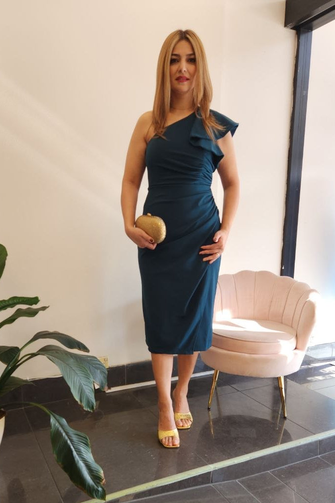 A woman stands indoors wearing a dark teal one-shoulder midi dress and holding a small round clutch. The setting includes a potted plant, a pale pink armchair, a window, and a mannequin displaying a light blue gown in the background.