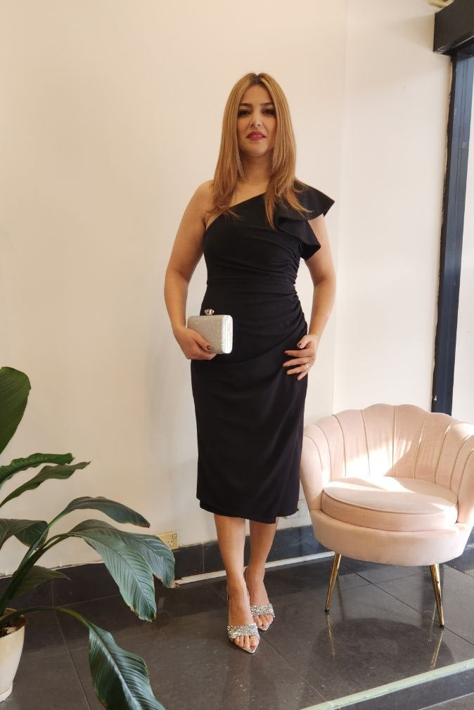 A woman stands indoors wearing a black one-shoulder midi dress and holding a small white cup. The setting includes a potted plant, a pale pink upholstered chair, and a dark tiled floor.