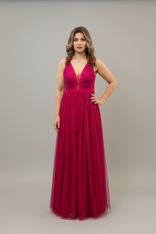 A woman modeling a floor-length magenta formal gown with a deep V-neckline, lace bodice, and flowing tulle skirt against a plain gray studio background.