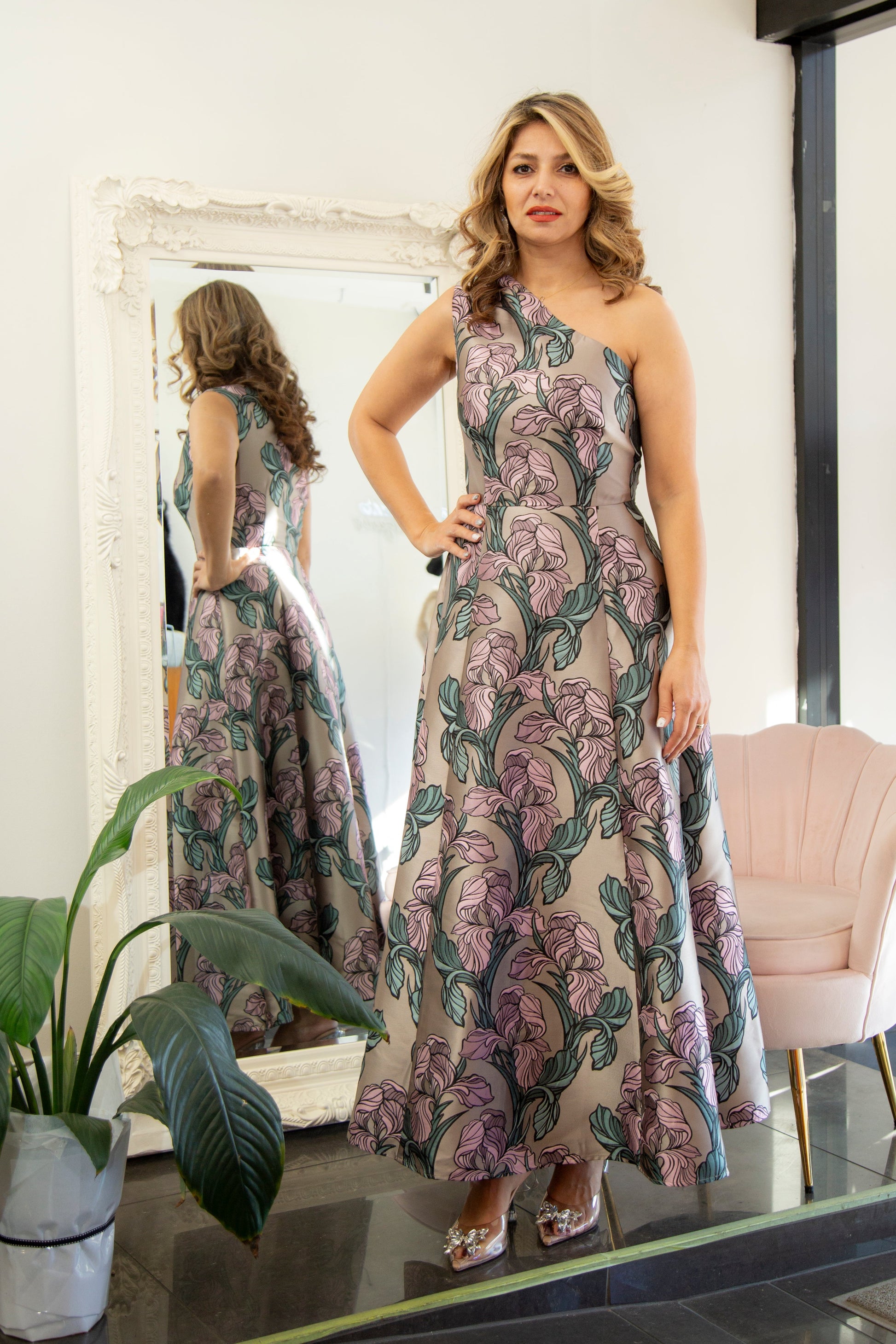 Woman in a floral dress standing in front of a mirror in a room with a plant and chair.