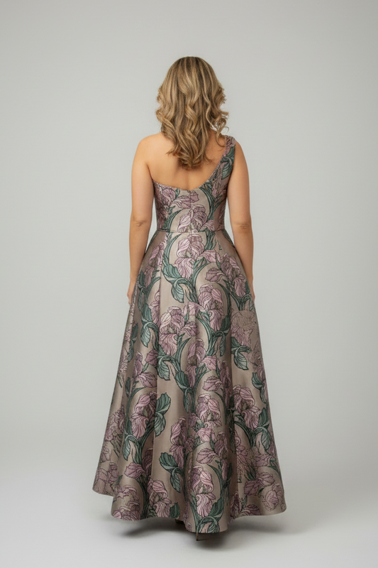 A woman viewed from behind wearing a sleeveless floor-length gown with a floral pattern in muted pink, green, and taupe tones against a light gray studio background.