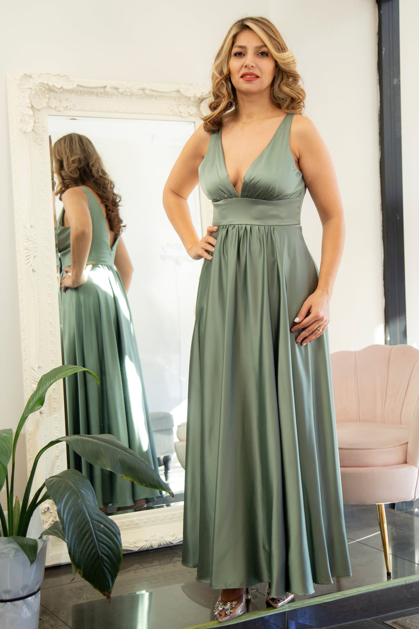 Woman in a green dress standing in front of a mirror in a room with a plant and chair.