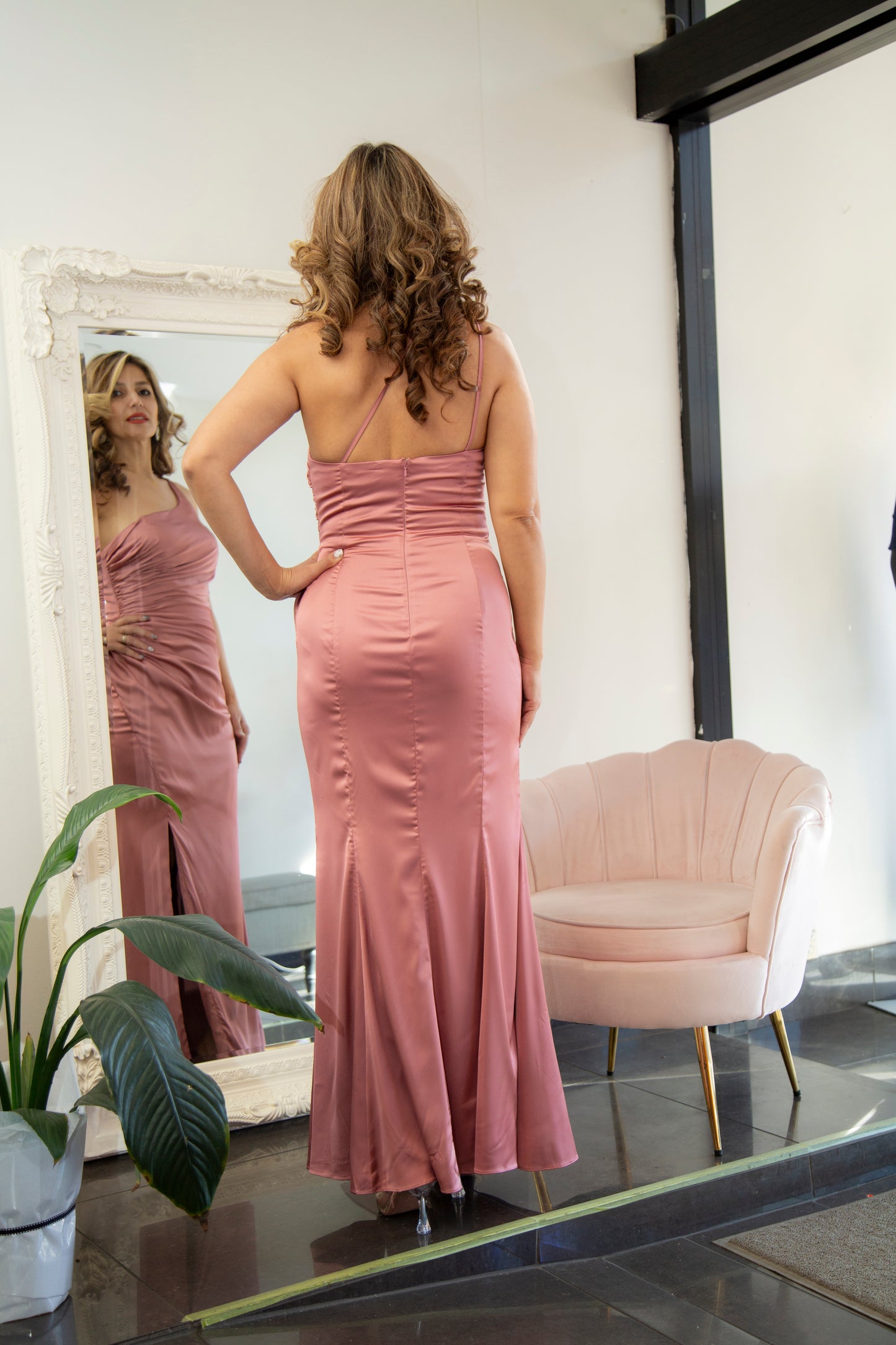 Woman in a pink dress standing in front of a mirror in a room with a chair and plant.